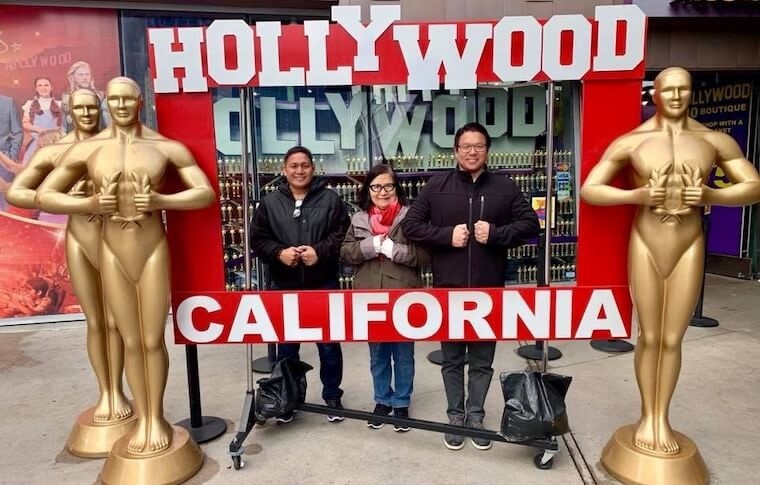 Three people pose under 'Hollywood California' sign with Oscar statues.