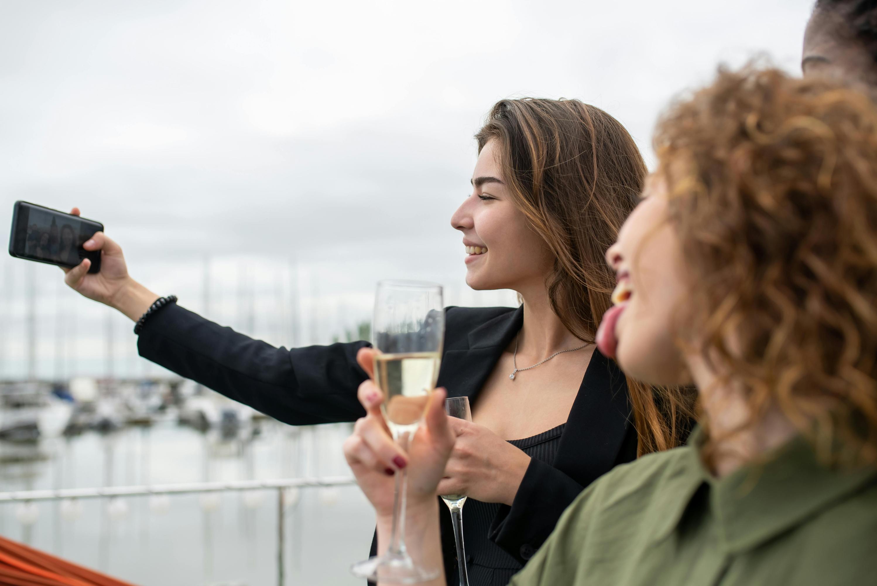 Two women taking a selfie with champagne glasses by a marina.