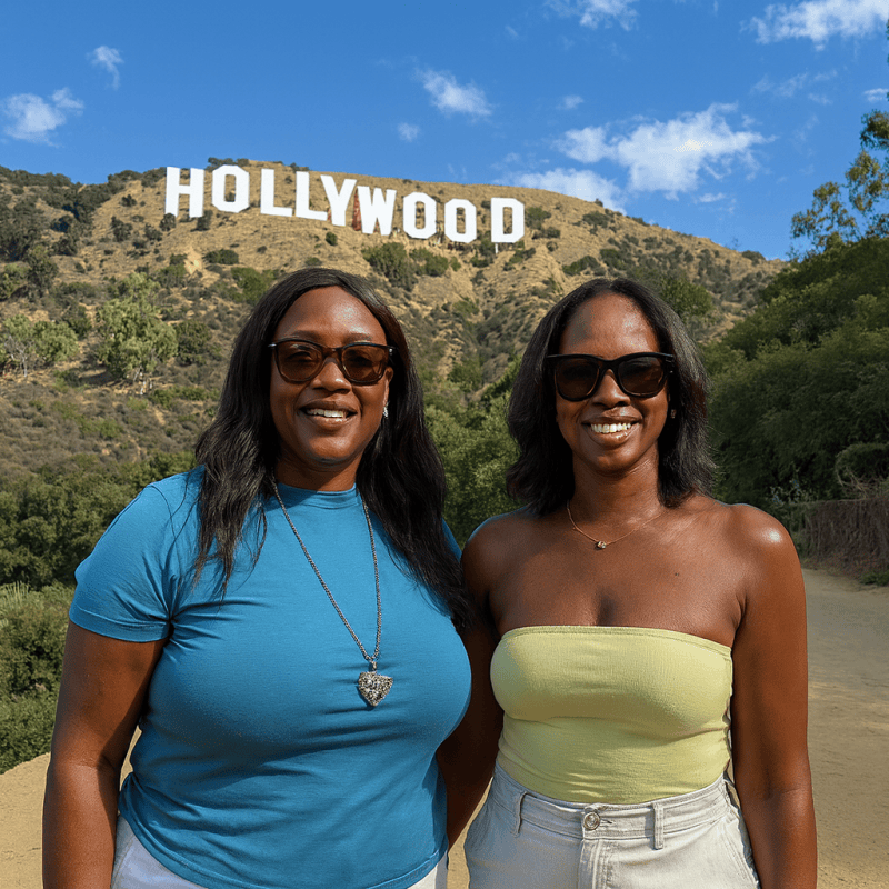 Two women from Nigeria smiling on the Hollywood Sign hiking trail with the iconic white letters visible on the hillside behind them