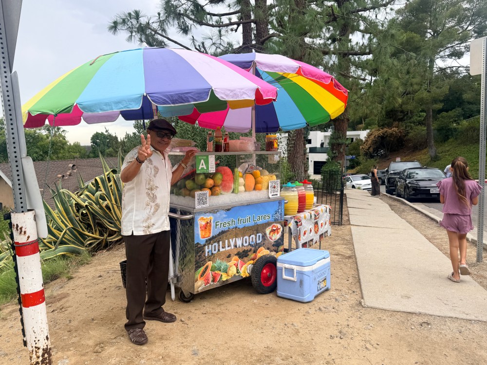 Tourist smiling and giving peace sign next to fruit vendor cart on Innsdale Trail in Lake Hollywood Estates